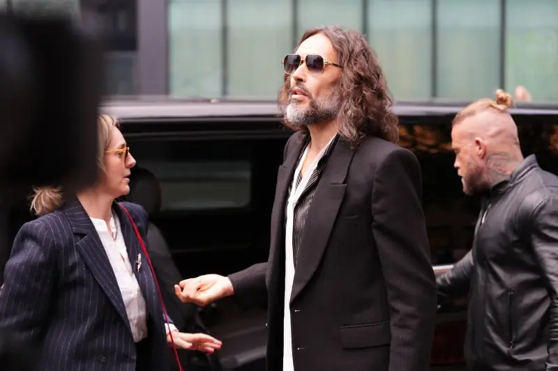 A man in a suit and sunglasses is seen interacting with another individual in a pinstriped suit, with a black vehicle and another person in the background.