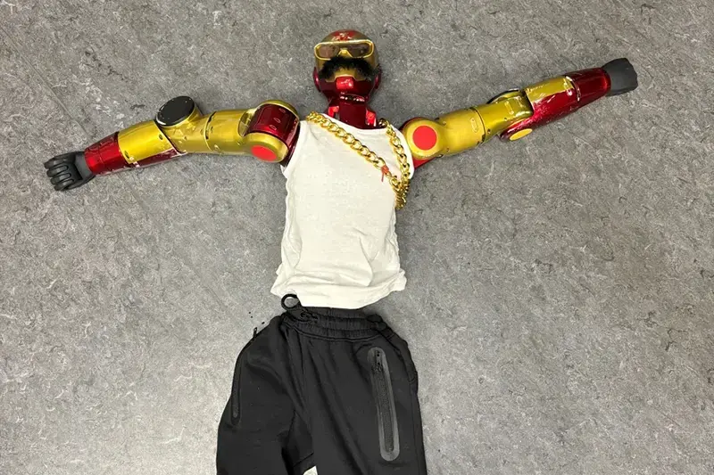 A person dressed in a DIY Iron Man costume lies on the ground with arms outstretched, wearing a white shirt, black pants, and a gold chain.