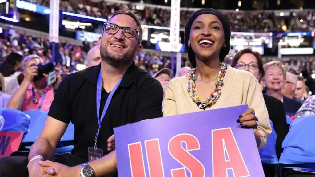 A man and a woman are seated in a crowd, smiling and holding a sign that reads "USA."