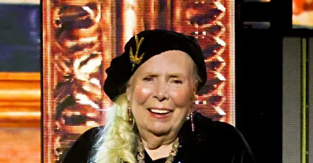 The image shows a person with long blonde hair styled in a braid, wearing a black outfit and a decorative headpiece, smiling in front of a backdrop with a golden design.