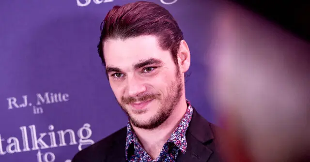 The image shows a man with a neatly styled hairstyle and a beard, wearing a patterned shirt and a suit jacket, smiling at the camera in front of a purple backdrop with text.
