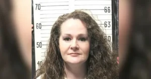 The image appears to be a mugshot of a woman with long, curly hair, standing in front of a height chart.