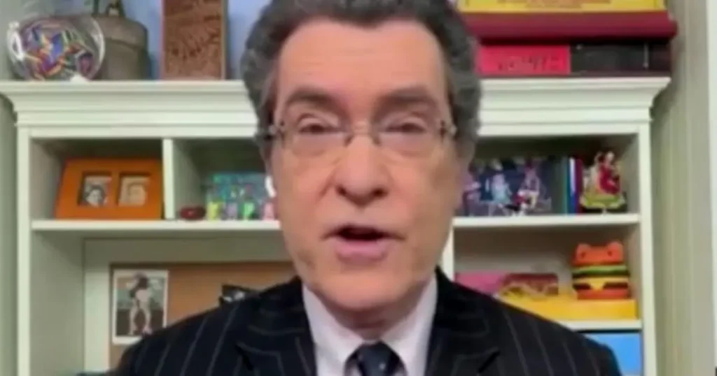 The image shows a person in a suit and tie, speaking in front of a bookshelf filled with various items and decorations.