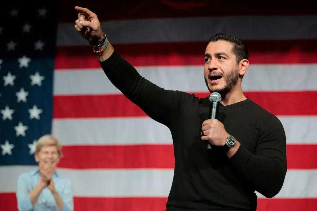A man speaks passionately into a microphone in front of an American flag, with a woman clapping in the background.