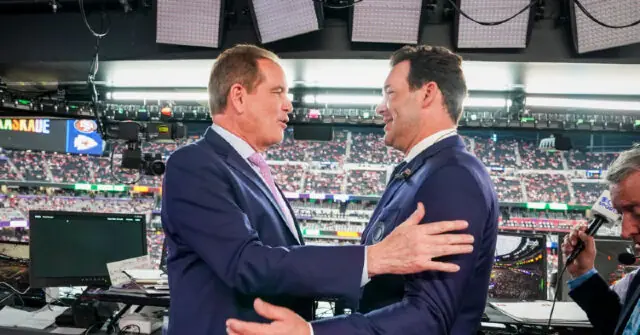 Two men in suits embrace in a broadcast booth at a large stadium filled with spectators.