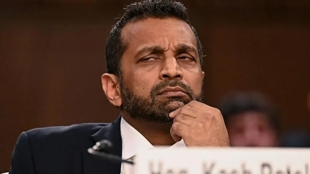 The image shows a man in a suit, appearing thoughtful, with his hand on his chin, seated at a table with a nameplate in front of him.