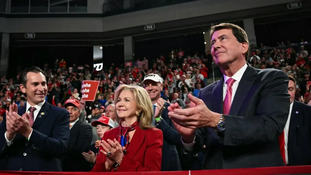A group of people, including a man in a suit and a woman in a red jacket, are clapping at a political rally.