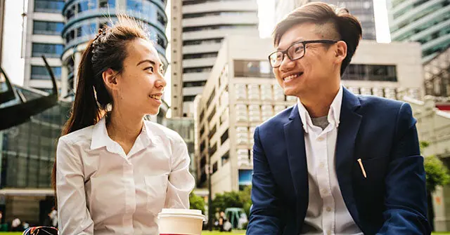 Two individuals are engaged in a conversation outdoors, with one holding a coffee cup, set against a backdrop of modern urban architecture.