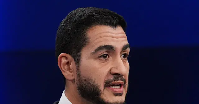 The image shows a man with a beard and short hair, wearing a suit, speaking in front of a blue background.