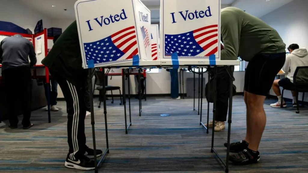 Two individuals are seen bending over at voting booths in a polling station, with "I Voted" signs prominently displayed.