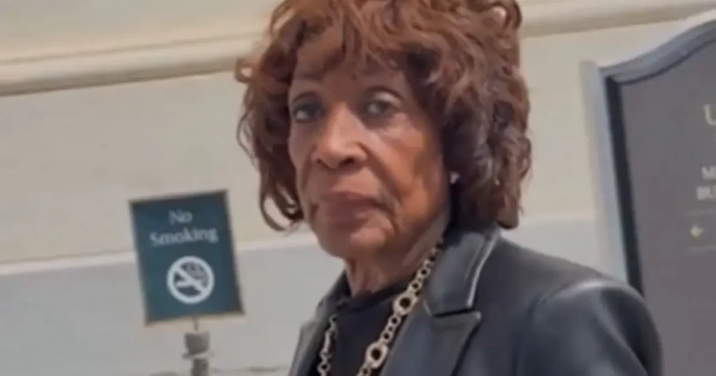 The image shows a person standing in front of a "No Smoking" sign, wearing a dark blazer and a necklace.