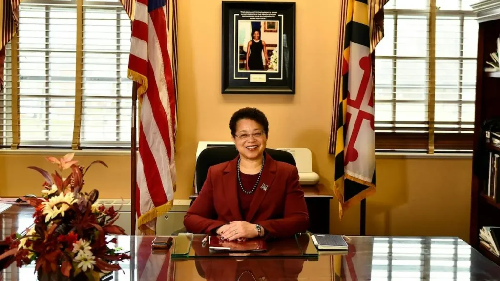 A woman in a professional setting smiles at the camera, seated at a desk with flags and a framed picture in the background.