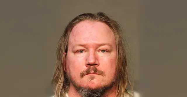 The image appears to be a mugshot of a man with long hair and a mustache, taken against a plain background.