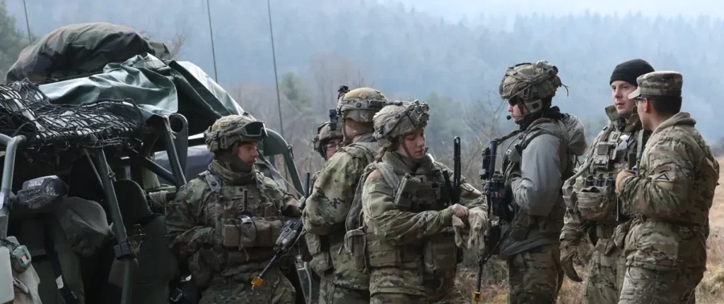 A group of soldiers in camouflage uniforms and tactical gear are gathered near a military vehicle, engaged in a discussion in an outdoor setting.