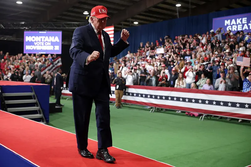The image shows a political rally in Montana, with a speaker on stage and a large audience in the background.