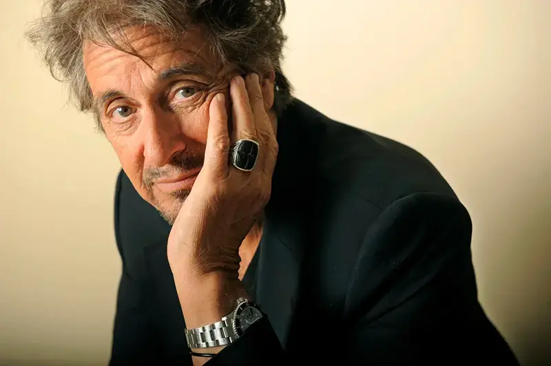 The image shows a person in a dark suit with a hand resting on their face, wearing a ring and a wristwatch.