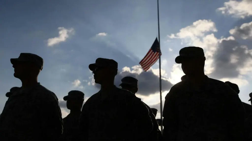 The silhouette of soldiers stands solemnly in front of a flag at half-staff, symbolizing respect and remembrance.