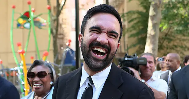 A man in a suit and tie is smiling widely in front of a crowd and colorful decorations.