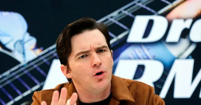 The image shows a person speaking in front of a backdrop featuring the "Drop the Mic" logo.