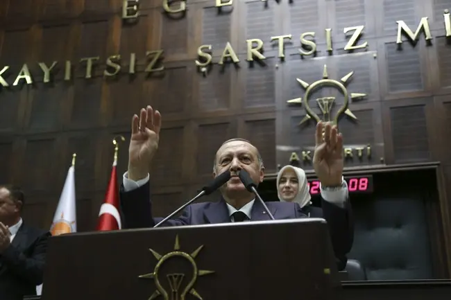 The image shows a person standing at a podium with their hands raised, in front of a backdrop featuring the text "KAYITSIZ ŞARTSIZ Mİ" and a logo, with flags and a clock visible in the background.