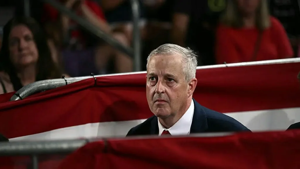 A man in a suit and tie is seated in a stadium, with an American flag partially visible in the foreground.