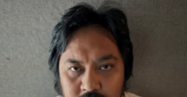 The image shows a close-up of a person with dark hair and a mustache, set against a plain background.