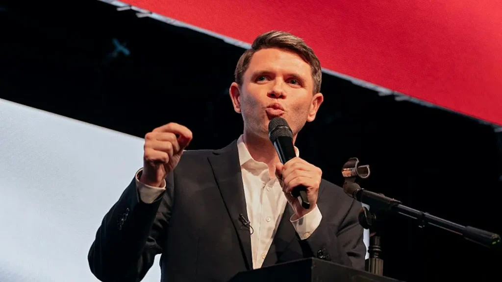 A man in a suit is speaking into a microphone at a podium, gesturing with his hand.
