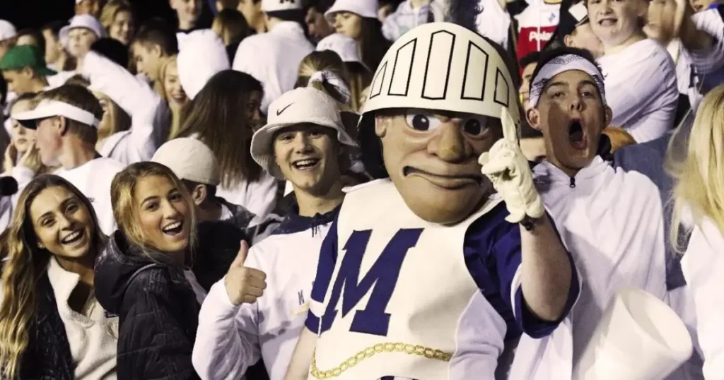 A group of enthusiastic fans and a mascot in a white and blue uniform celebrate at a sports event.