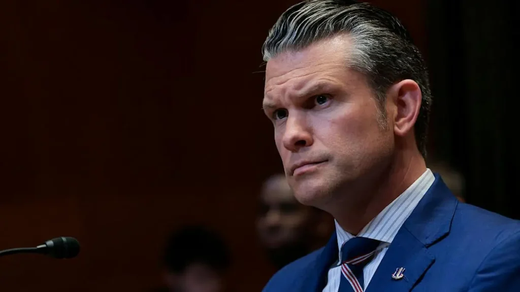 The image shows a man in a suit and tie, appearing to be in a formal setting, possibly giving a speech or participating in a discussion.