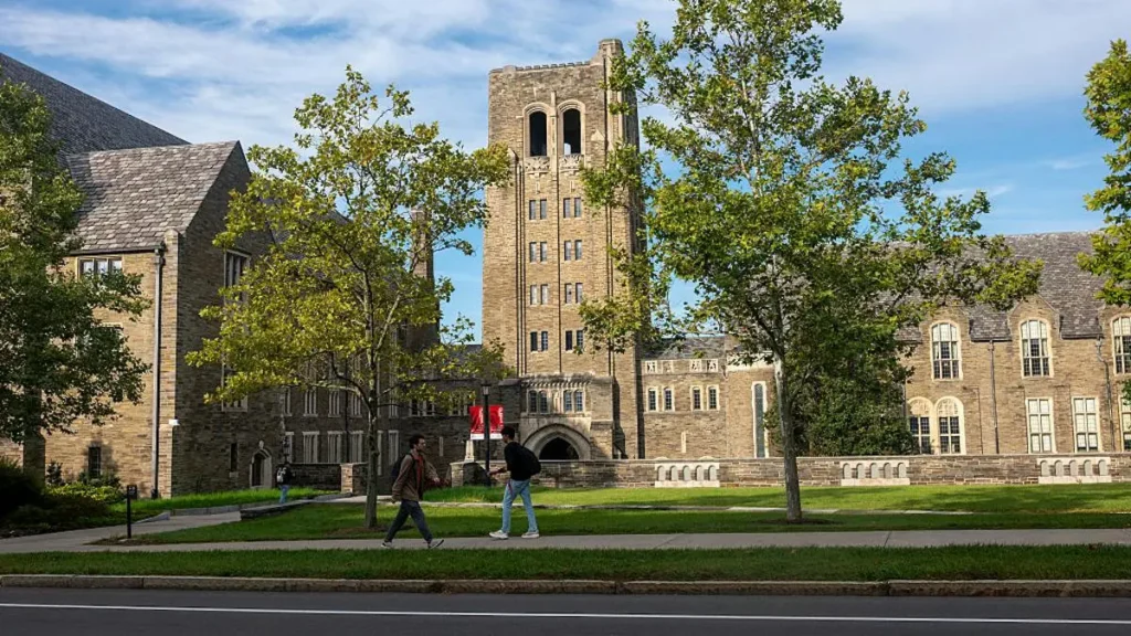 The image shows a picturesque campus scene with students walking on a sidewalk in front of a large, historic stone building with a tall tower, under a clear blue sky.