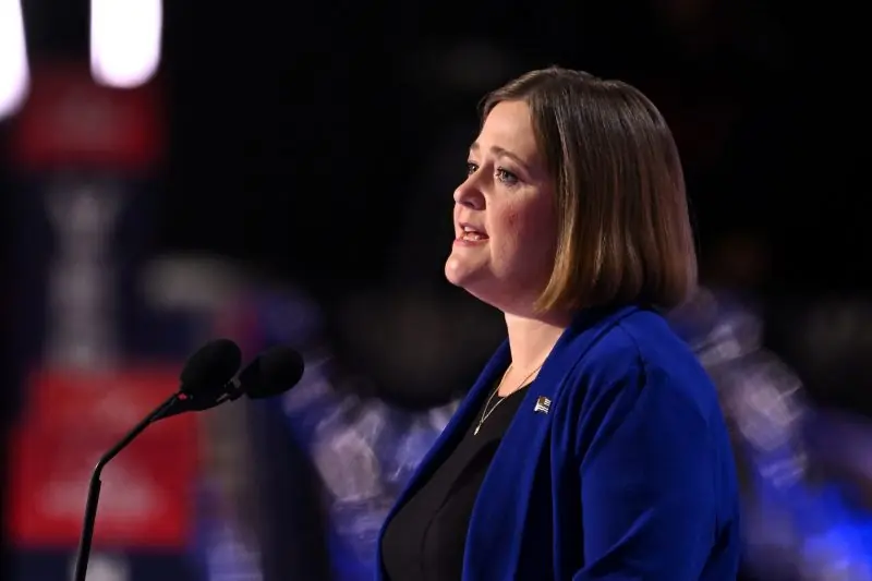 A woman in a blue blazer is speaking at a podium in front of a blurred audience.
