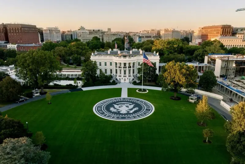 The image shows an aerial view of the White House with the presidential seal prominently displayed on the lawn in front of it.