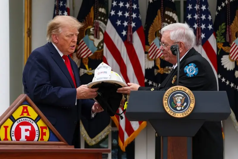 The image shows a formal event where a person is presenting a firefighter's helmet to another individual at a podium with the seal of the President of the United States in the background.