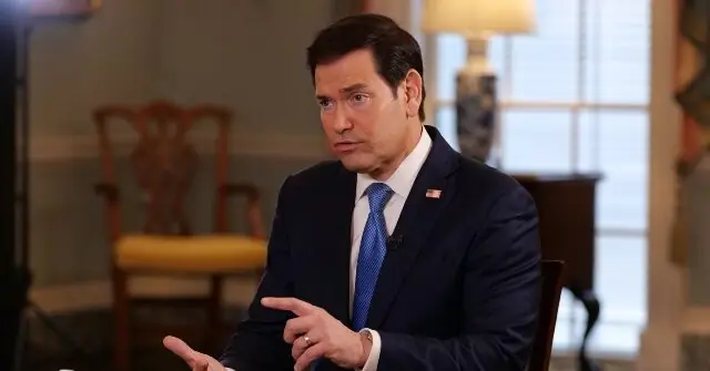 The image shows a man in a suit and tie, seated and gesturing with his hands, likely during an interview or news segment.