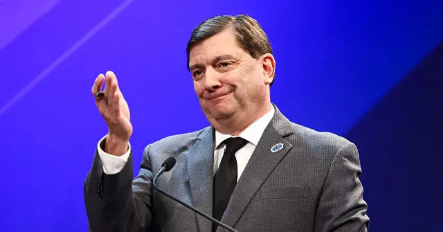 The image shows a man in a suit gesturing with his right hand while speaking at a podium, set against a blue background.