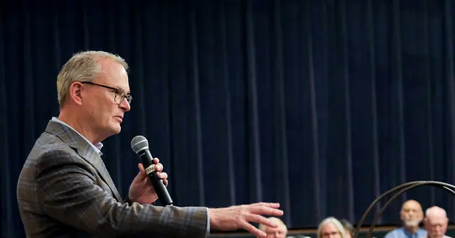 A man in a suit is speaking into a microphone while gesturing with his other hand, with an audience seated in the background.