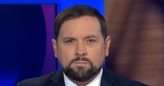 The image shows a man in a suit and tie, appearing on a news broadcast with a serious expression.