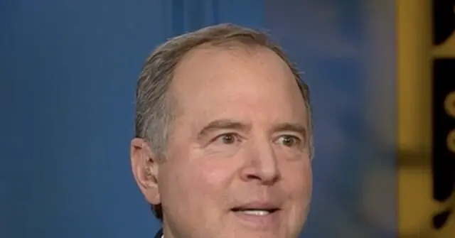 The image shows a close-up of a man speaking, with a blurred background that includes a blue and yellow element.