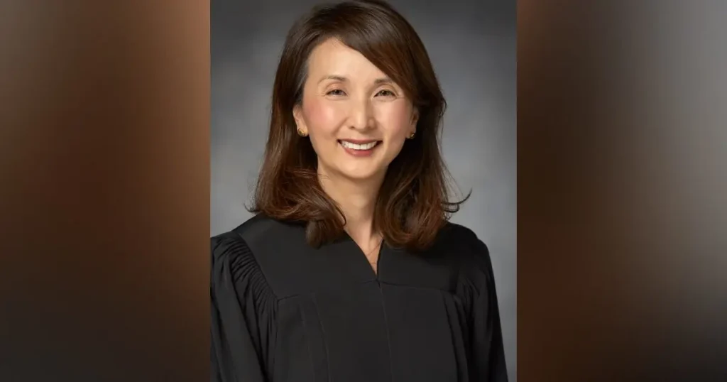 The image shows a person wearing a black robe, likely a judge or academic, against a neutral background.