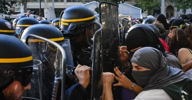 The image depicts a tense confrontation between police in riot gear and protesters during a demonstration.