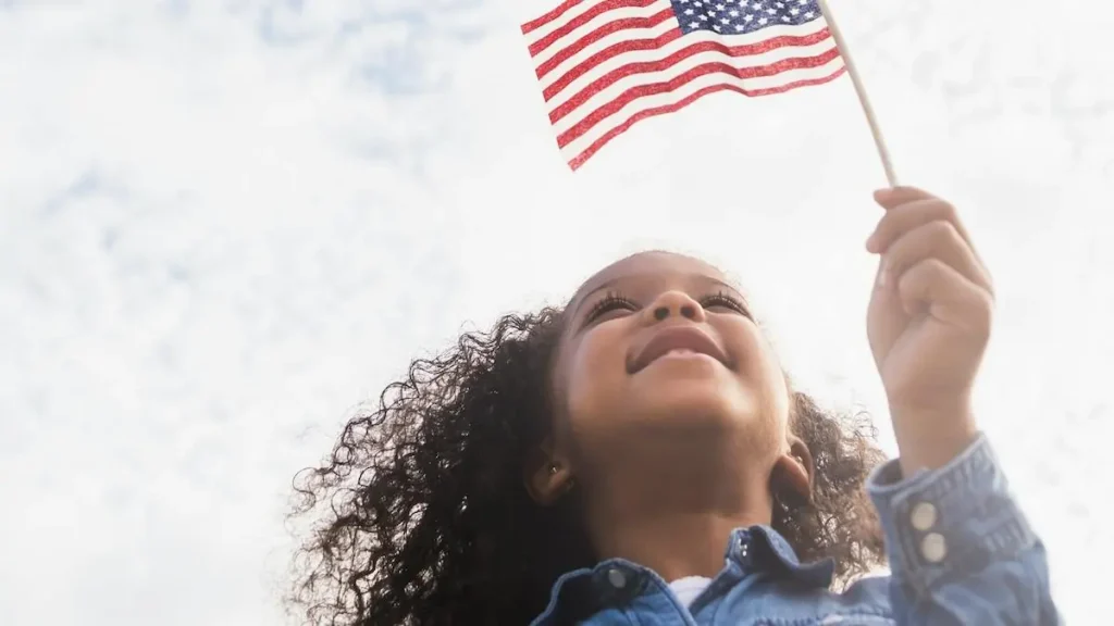 A young child holds an American flag, smiling and looking up, symbolizing patriotism and national pride.