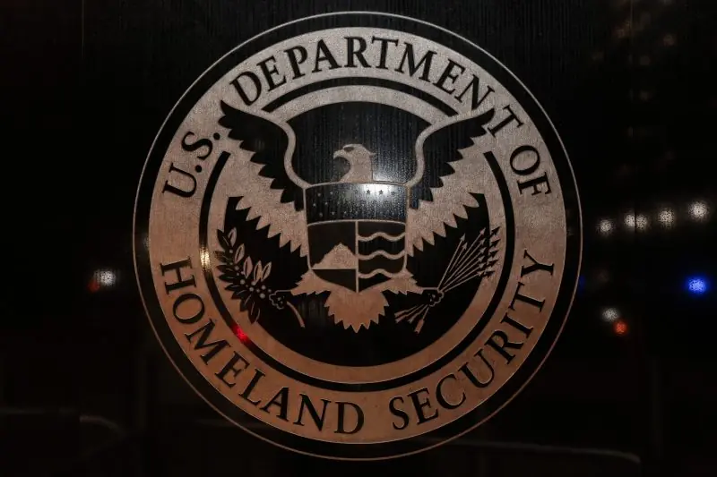 The image shows the seal of the U.S. Department of Homeland Security, featuring an eagle and shield, set against a dark background with some blurred lights.