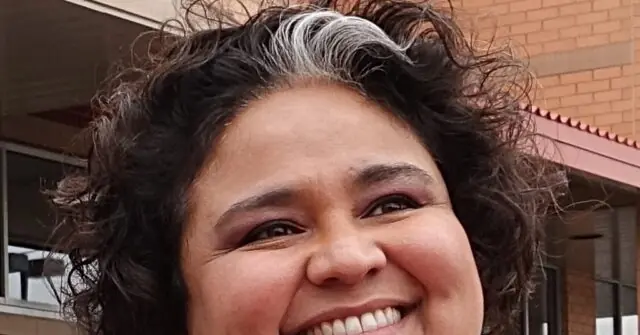 The photo shows a person with curly hair smiling in front of a brick building.