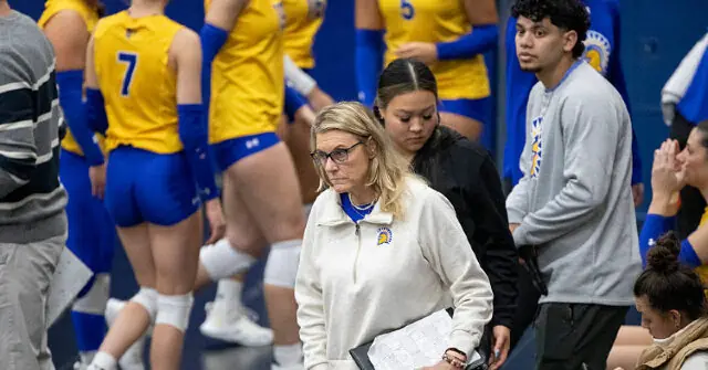 A volleyball coach and players are seen on the sidelines during a game.