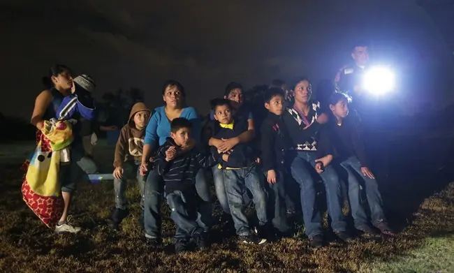 A group of people, including children, are gathered outdoors at night, illuminated by a flashlight, suggesting a moment of shared experience or concern.