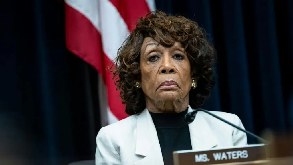 The image shows a person seated at a podium with a nameplate that reads "Ms. Waters," with an American flag in the background.