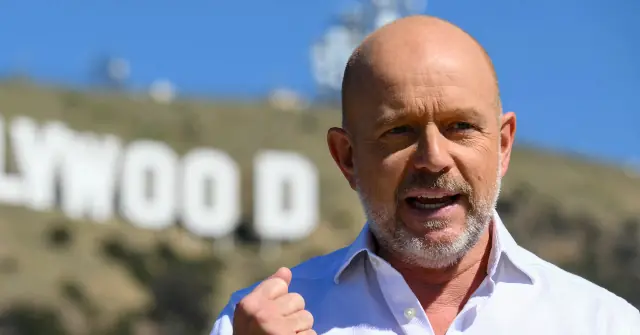 A man stands in front of the Hollywood sign, gesturing with his fist.
