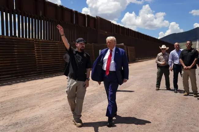 The photo shows a group of individuals, including a man in a suit, walking along a border wall under a clear sky.