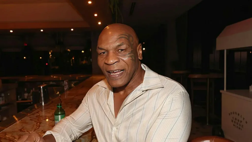 The image shows a man sitting at a bar, smiling, with a bottle of green liquid on the counter in front of him.