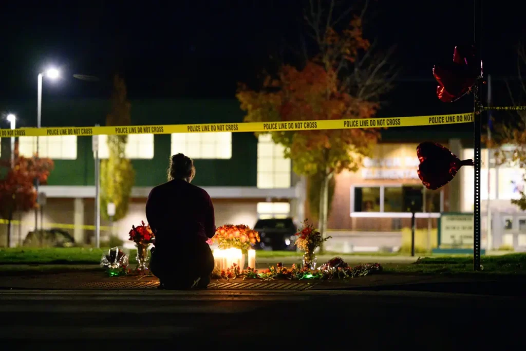 A person sits in front of a makeshift memorial with candles and flowers, surrounded by police tape at night.